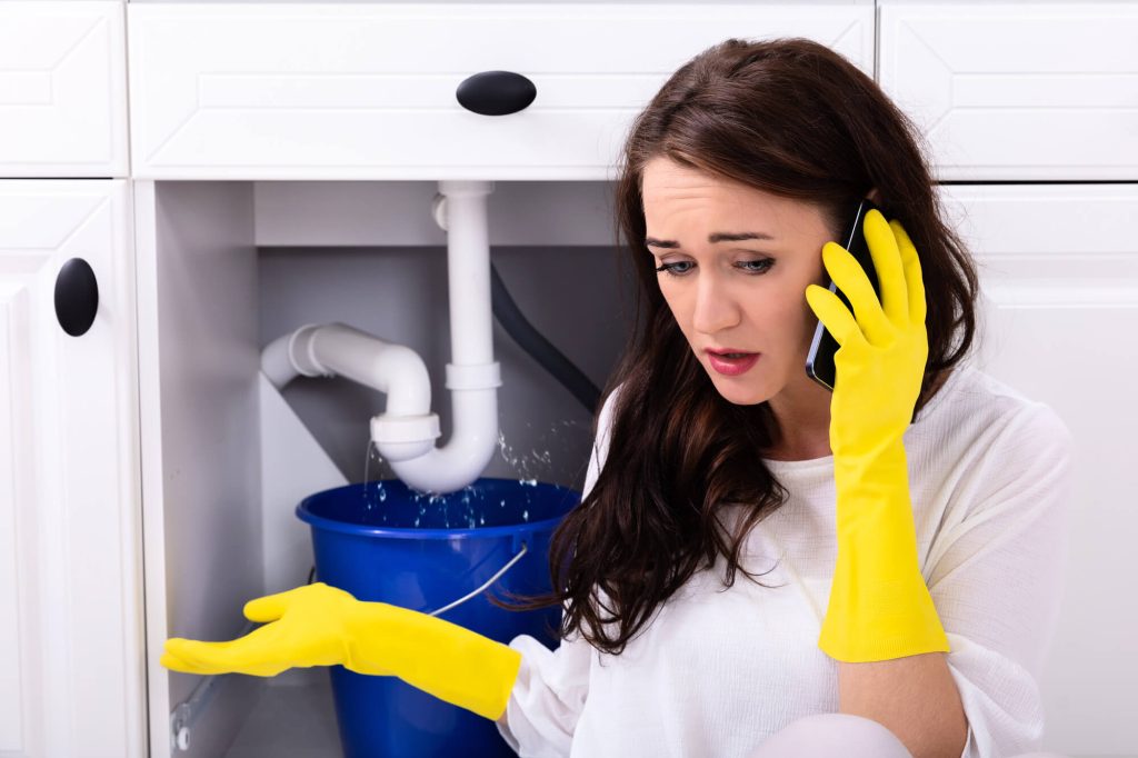 Sad Young Woman Calling Plumber In Front Of Water Leaking from kitchen sink